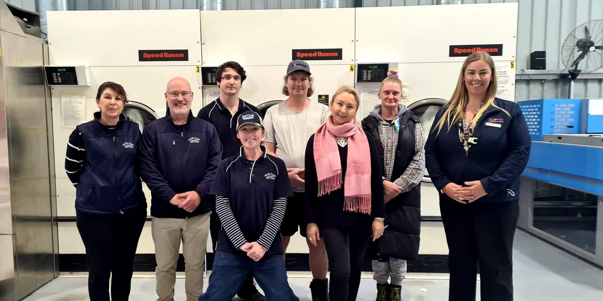 A group of employees from South Coast Laundry Services stand in the work area near some machinery